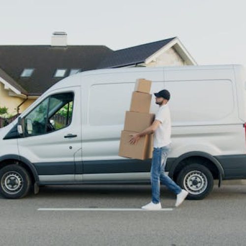 Man unloading cardboard boxes from a delivery van in a suburban neighborhood street.